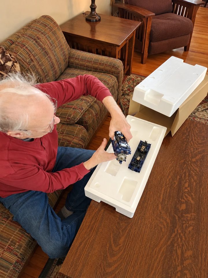 A collector on a couch lifting a blue locomotive out of its foam packing, its tender still in the box